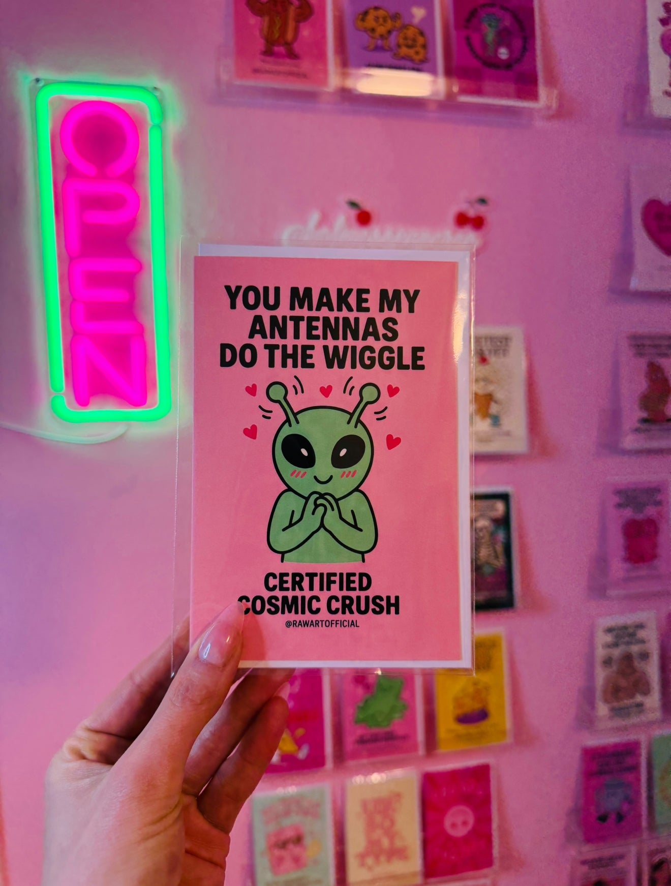 Cute green alien blushing with wiggling antennas and hearts on pink background, valentine card art.