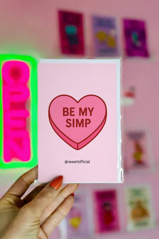 Funny greeting card with a pink candy conversation heart reading “Be My Simp” on a light pink background.
