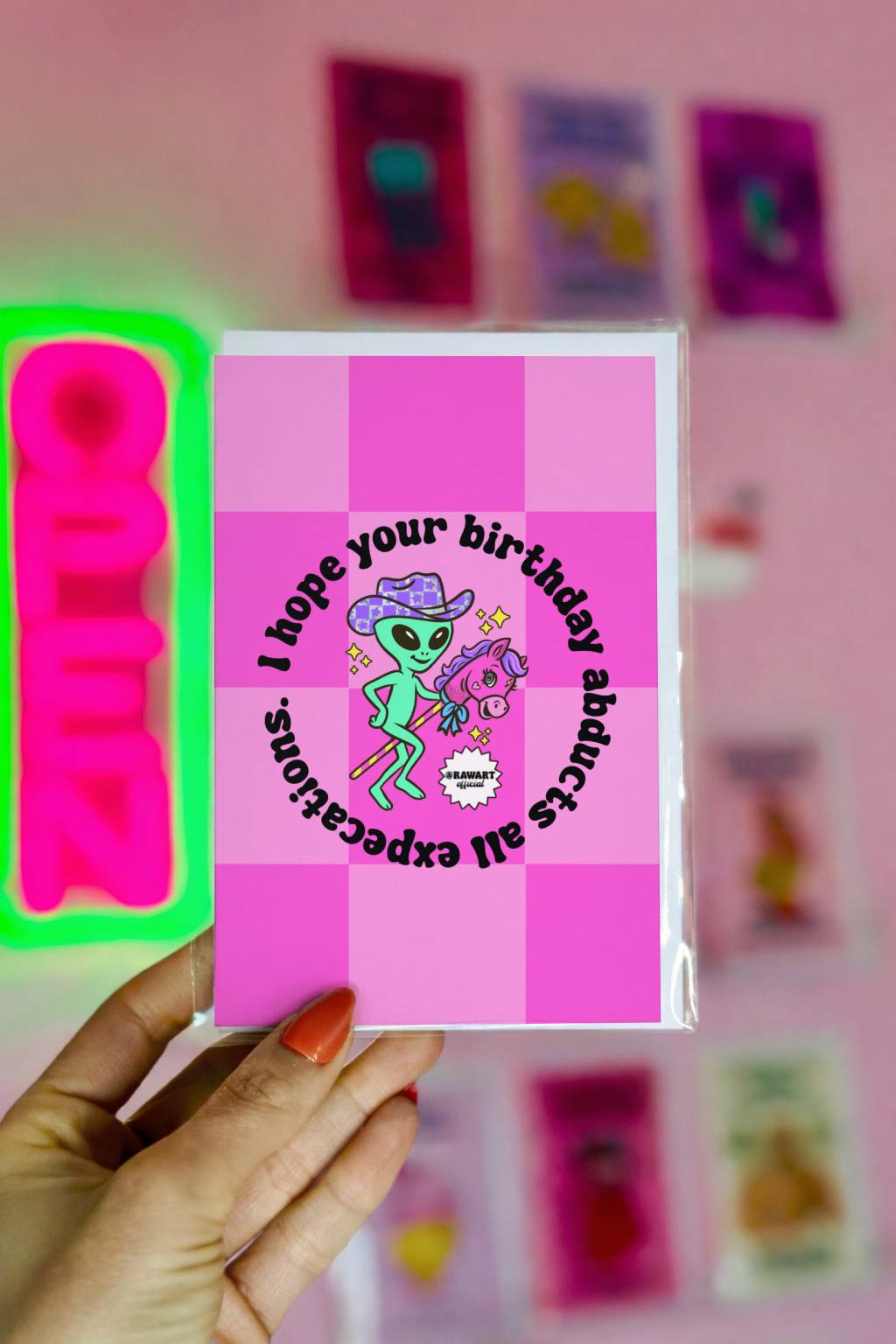 Pink checkerboard birthday card with green alien in cowboy hat riding stick horse under circular text about birthday abduction.