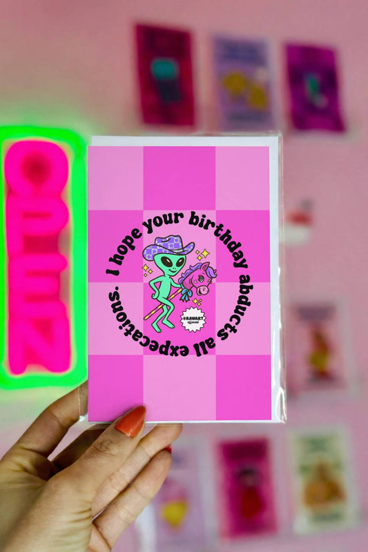 Pink checkerboard birthday card with green alien in cowboy hat riding stick horse under circular text about birthday abduction.