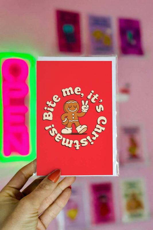Funny Christmas greeting card featuring a retro gingerbread character making a peace sign with circular text reading “Bite Me It’s Christmas.”