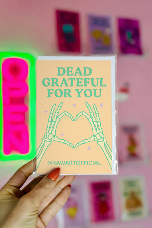 Card with 'Dead Grateful For You' text held by a hand against a pink background with colorful cards.