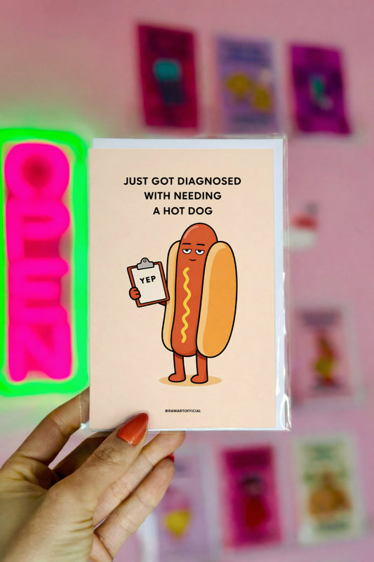 Card with a cartoon hot dog holding a sign saying 'Just got diagnosed with needing a hot dog' against a pink background with colorful cards.