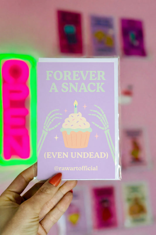 Halloween card with skeleton hands holding cupcake and text “Forever a Snack (Even Undead)” in pastel purple and green.