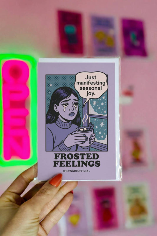 Funny Christmas greeting card featuring a retro comic-style illustration of a woman holding hot cocoa by a snowy window with the text “Frosted Feelings.”