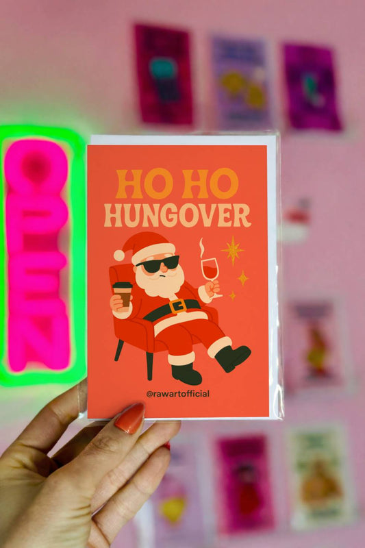 Funny Christmas greeting card featuring Santa relaxing in a chair with coffee and wine under the words “Ho Ho Hungover.”