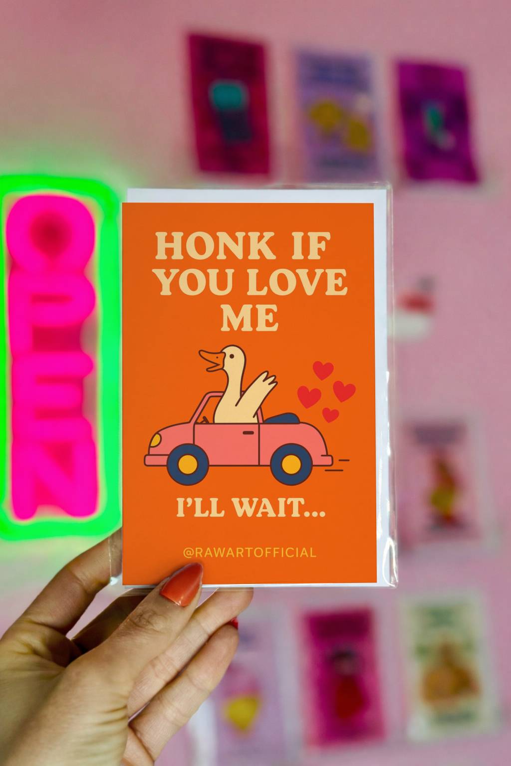 Bright orange greeting card with duck driving pink car and text Honk If You Love Me I’ll Wait and @rawartofficial at bottom.