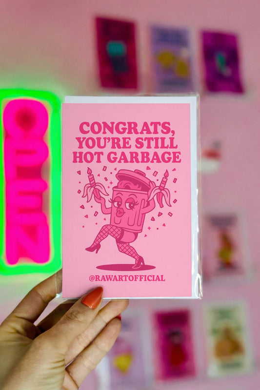 Pink retro-style greeting card with a dancing trash can holding candles and text “Congrats you’re still hot garbage” in bold lettering