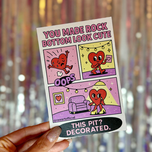 Funny pastel greeting card with comic panels of a heart character falling, decorating their emotional pit with string lights and a framed photo. Text reads: “You made rock bottom look cute. This pit? Decorated.” Designed in Y2K retro style with @rawartofficial branding.