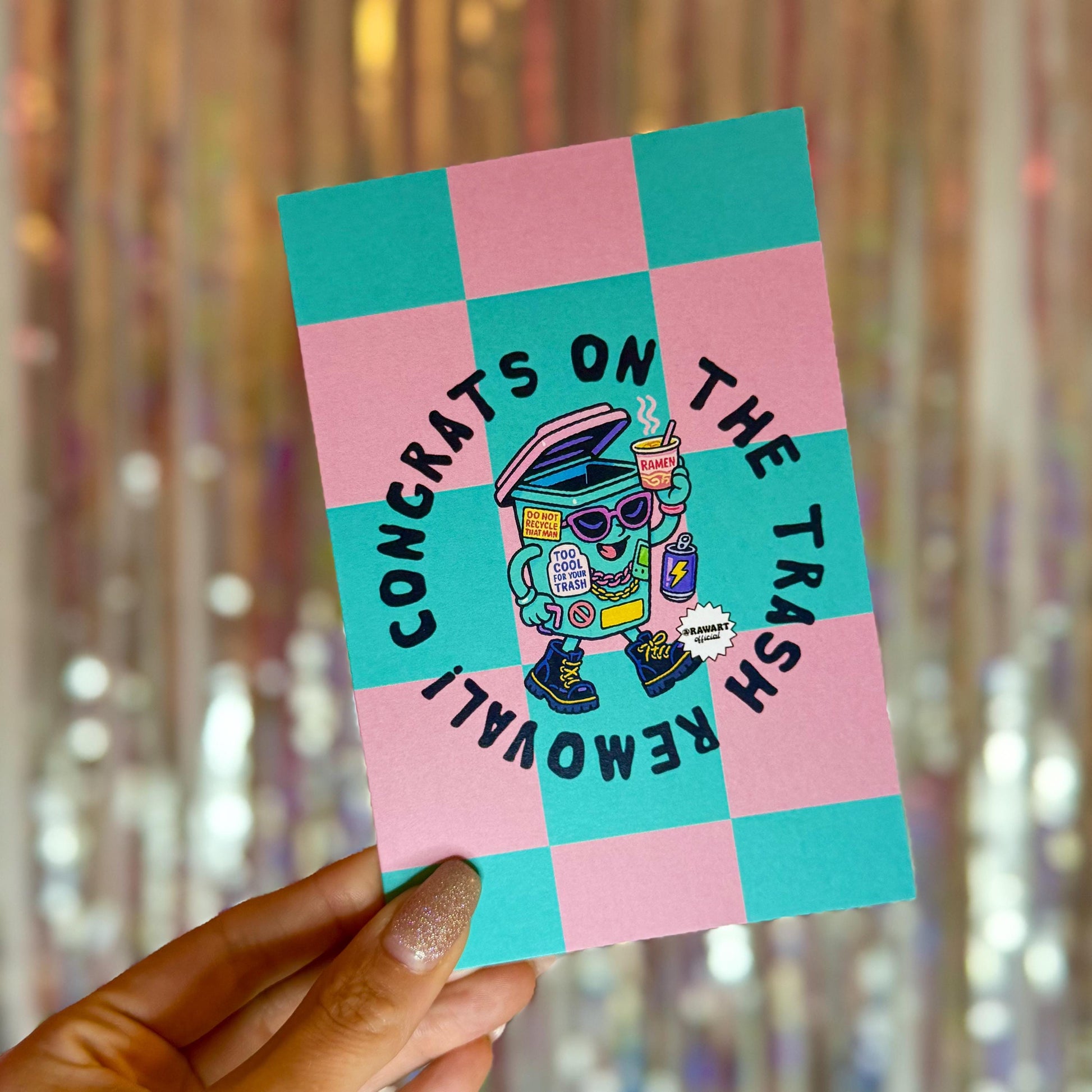 Front of pink and teal checkered breakup card featuring a walking trash bin character with sunglasses, boots, and stickers. Text reads “Congrats on the Trash Removal!” with a cheeky RAW Art Official logo.