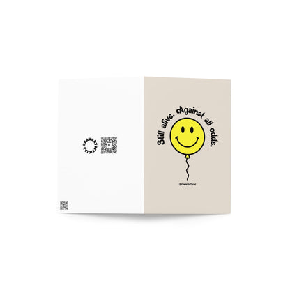 Retro yellow smiley balloon with text “Still alive. Against all odds.” on beige background