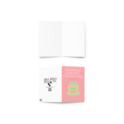 Pink greeting card that reads ‘CANDLE COUNT: TERRIFYING’ above a melting skull-faced birthday cake with many lit mint candles and sparkles—spooky, pastel-goth vibe.