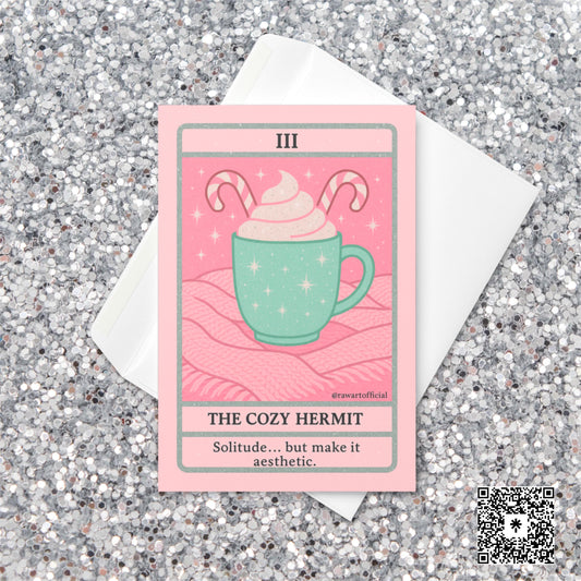 Pastel pink and mint Christmas tarot-style card featuring a mint green mug topped with whipped cream and candy canes on a pink knit blanket, titled “The Cozy Hermit” with the phrase “Solitude… but make it aesthetic.”