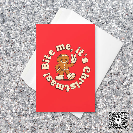 Red Christmas card featuring a smiling gingerbread man with a bite out of his leg, holding up a peace sign, surrounded by cream text reading “Bite me, it’s Christmas!”