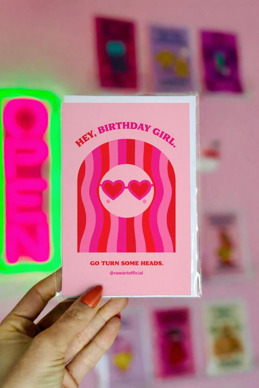Pink birthday card featuring a girl with wavy red and pink hair and heart-shaped sunglasses with the phrase “Hey, Birthday Girl. Go Turn Some Heads.”