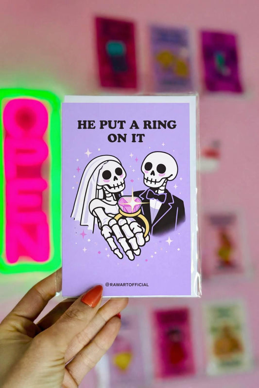 Lavender wedding card featuring skeleton bride and groom holding a large pink gemstone ring with text “He Put a Ring On It.”
