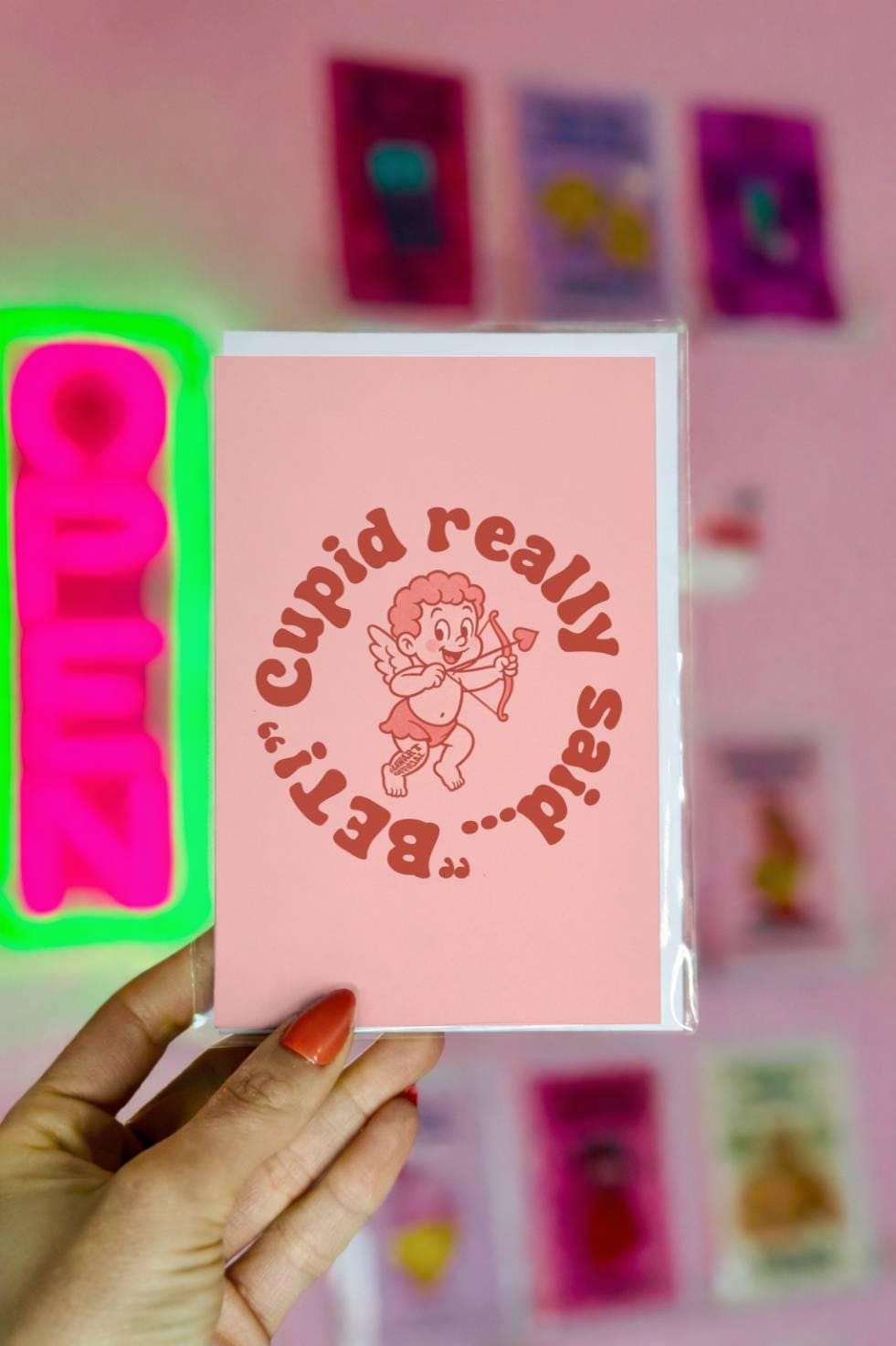 Funny wedding greeting card reading “Cupid Really Said Bet” with retro cupid illustration on a soft pink background.