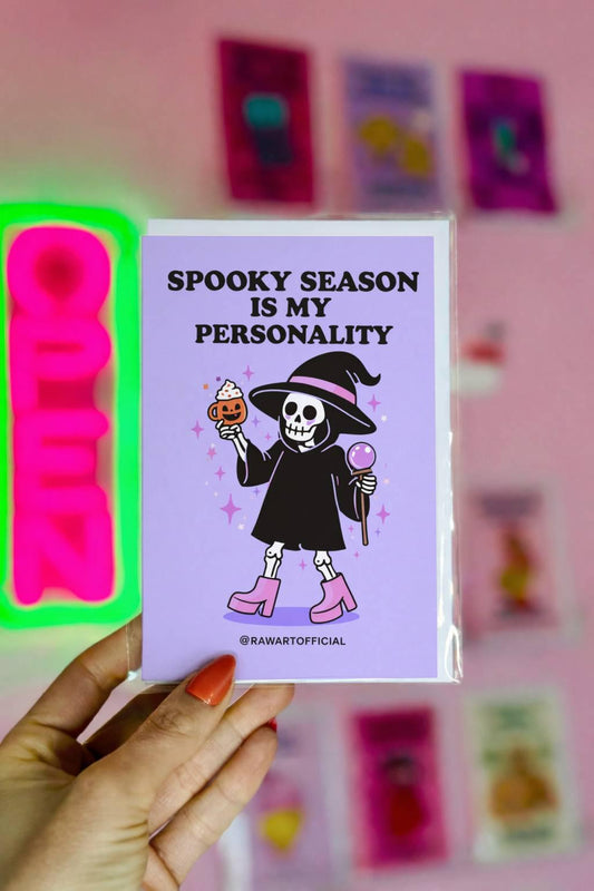 Spooky greeting card with skeleton in witch hat holding pumpkin drink and crystal ball with text “Spooky season is my personality” on purple background