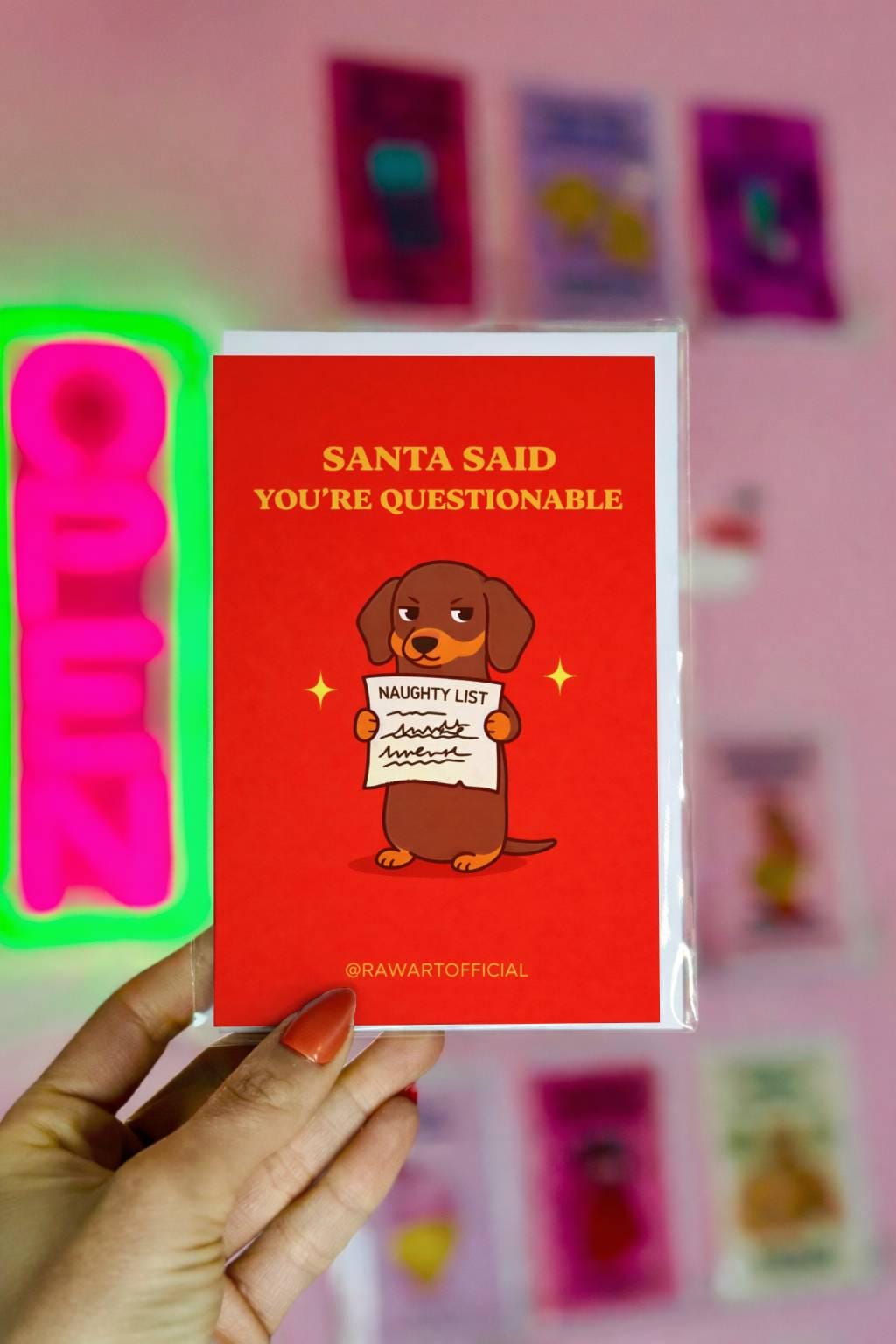 Funny dachshund Christmas greeting card with text “Santa said you’re questionable” and wiener dog holding naughty list on red background