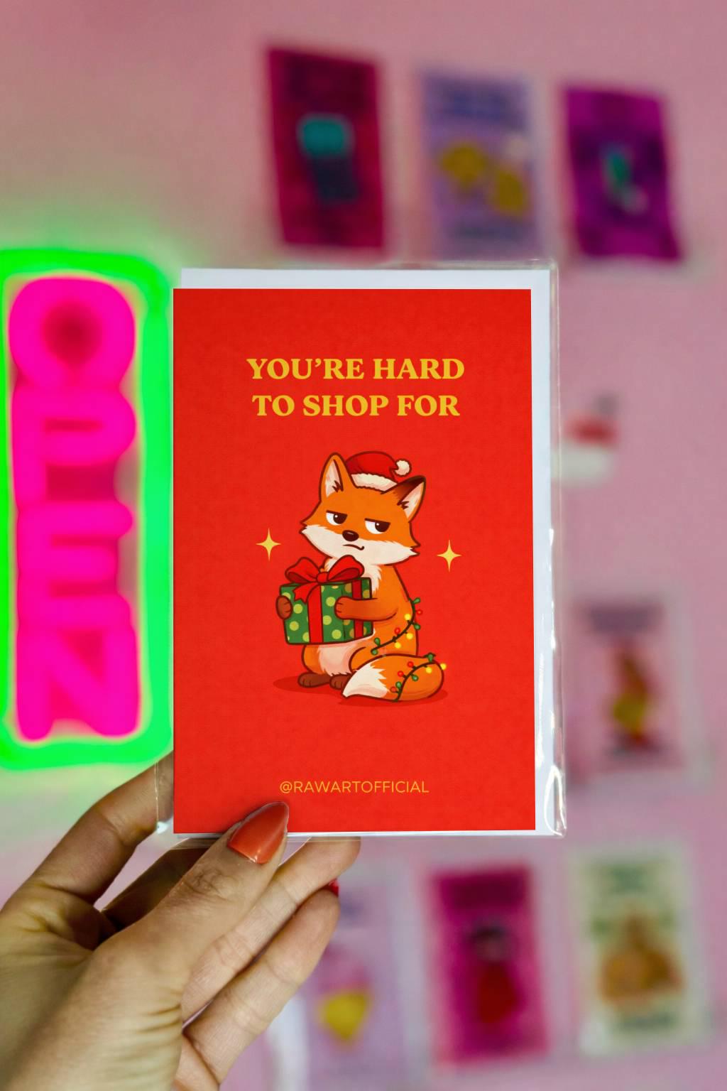 Funny Christmas greeting card with text “You’re hard to shop for” and fox holding gift illustration on red background