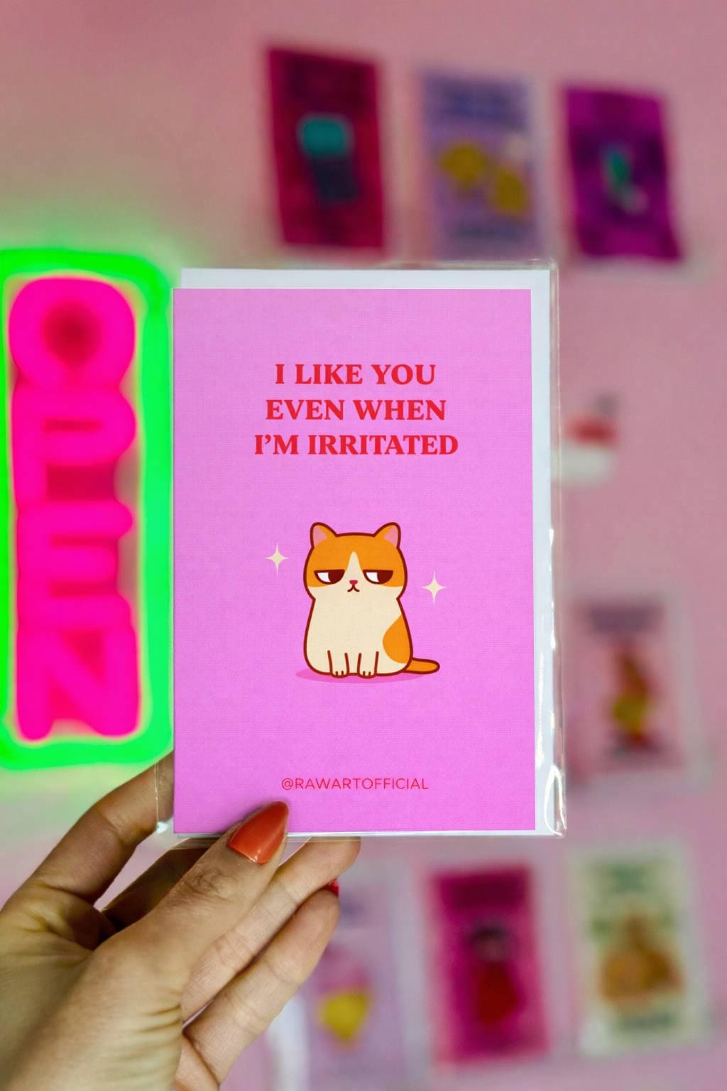 Funny love card featuring an unimpressed orange and white cat with text “I like you even when I’m irritated” on a pink background