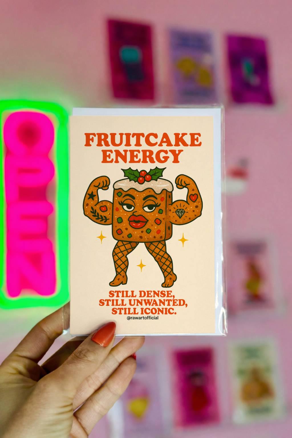 Funny Christmas card with muscular fruitcake character and text “fruitcake energy still dense still unwanted still iconic” on cream background