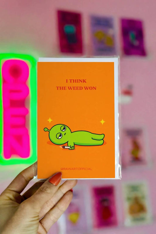 Funny stoner greeting card with green alien lying down holding joint, text says “I think the weed won” on orange background