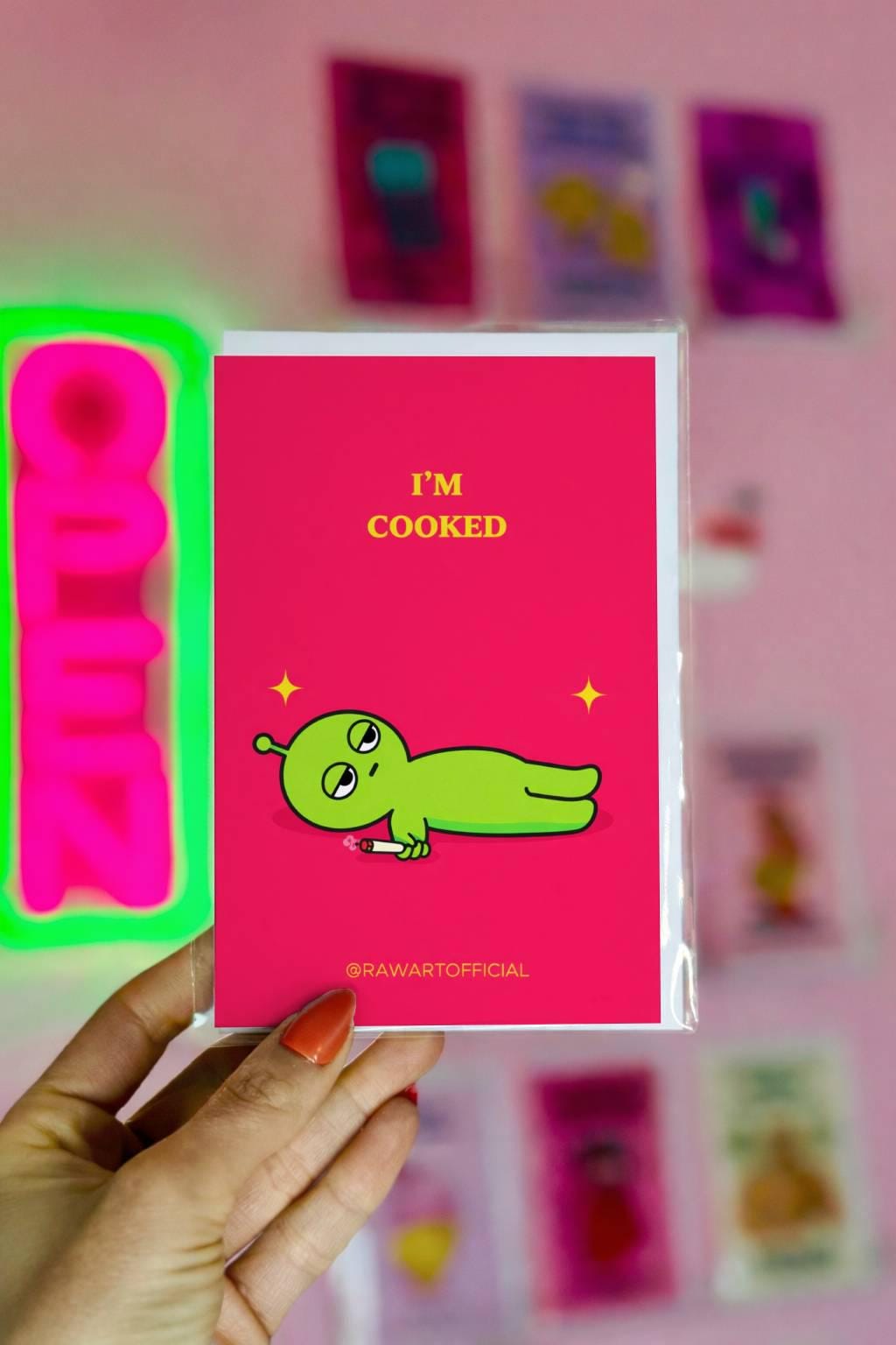 Funny stoner greeting card with green alien lying down holding joint, text says “I’m cooked” on pink background