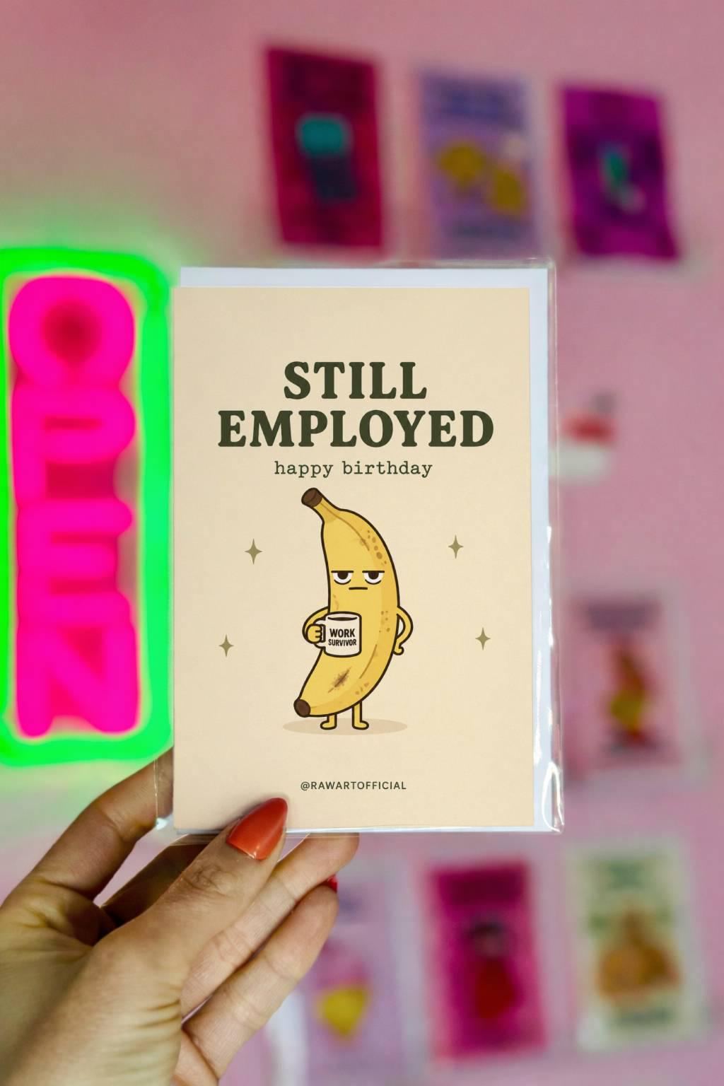 Funny coworker birthday greeting card with tired banana holding mug that says work survivor, text reads “Still employed happy birthday”