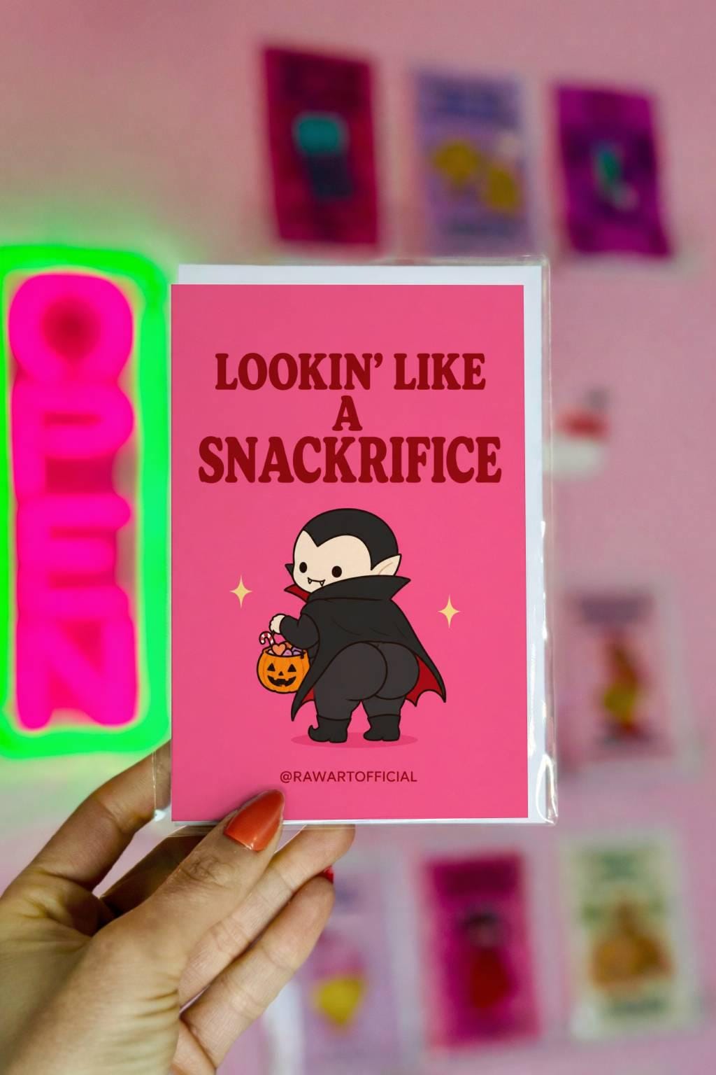 Funny Halloween greeting card with cute vampire booty holding pumpkin candy bucket on pink background saying “Lookin’ Like a Snackrifice”