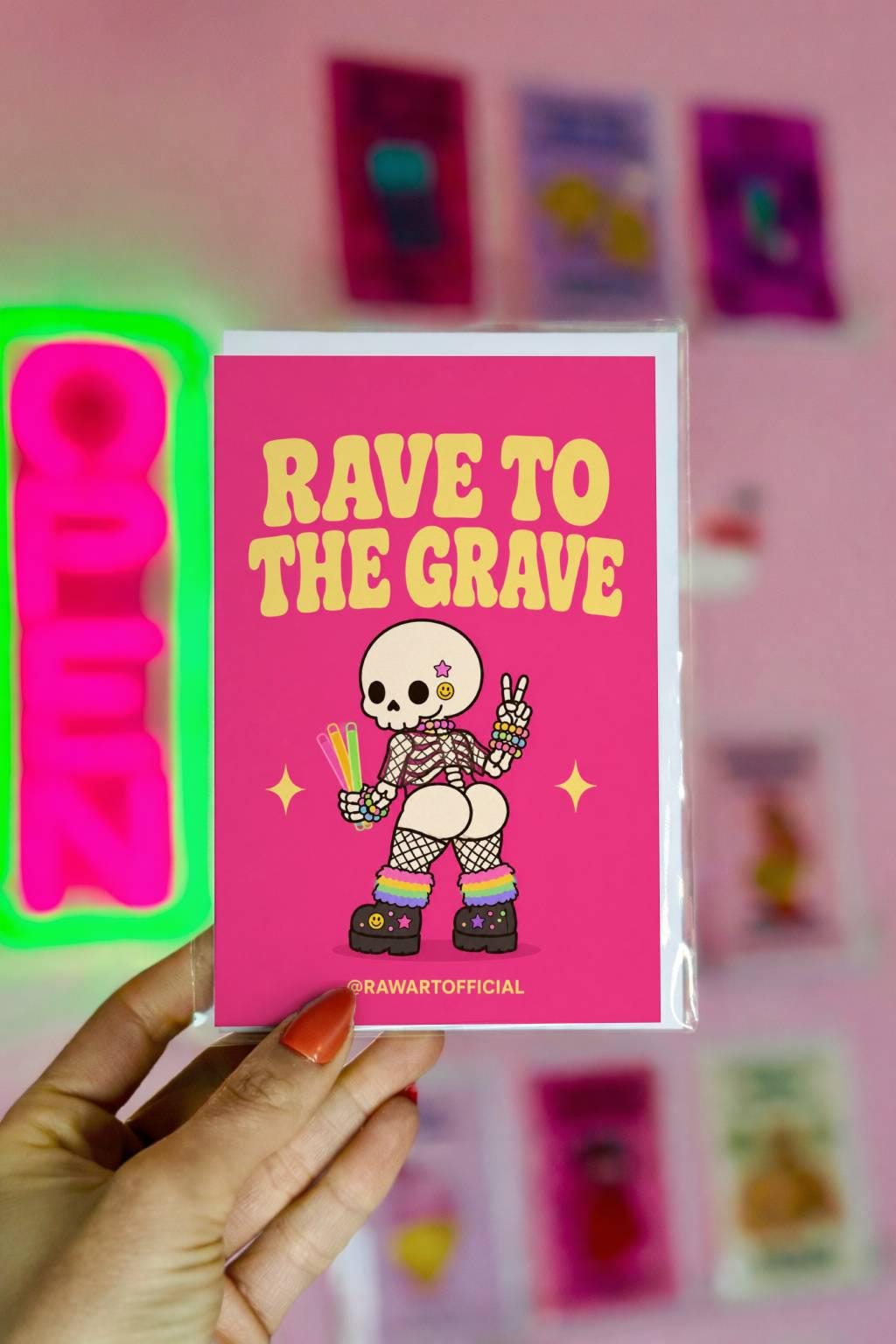 Funny Halloween greeting card with cheeked-up rave skeleton holding glow sticks and text reading “Rave to the Grave” on hot pink background.