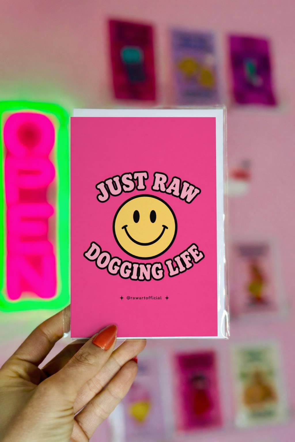 Funny pink greeting card with a retro smiley face and the text “Just Raw Dogging Life” in bubble lettering.