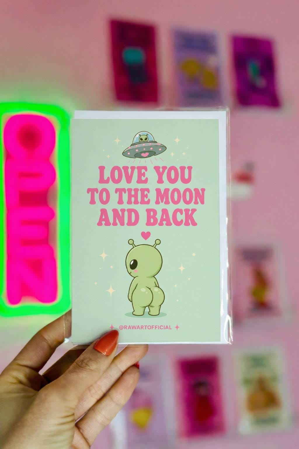 Funny love card featuring a cheeked-up alien under a UFO beside the phrase “Love You To The Moon And Back.”