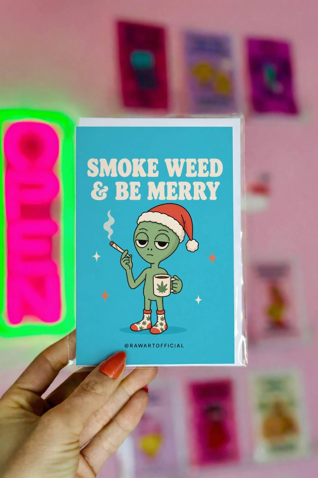 Funny Christmas card featuring an alien in a Santa hat holding a joint and weed mug beside the phrase “Smoke Weed & Be Merry.”
