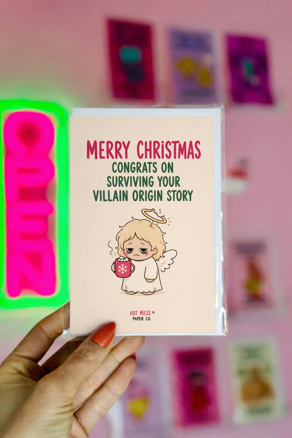 Funny Christmas card featuring a tired angel holding hot cocoa beside the phrase “Congrats On Surviving Your Villain Origin Story.”