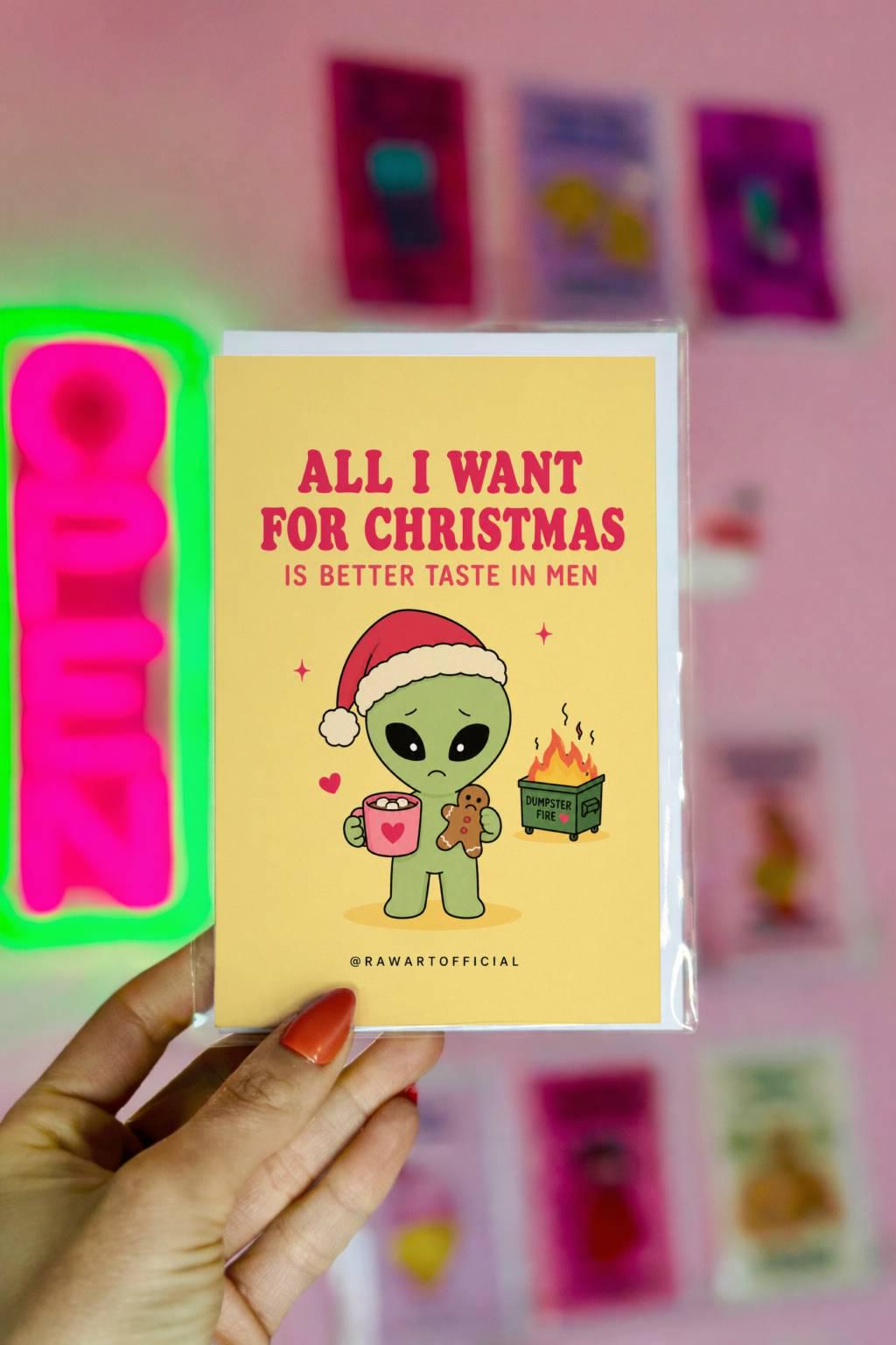 Funny Christmas card featuring a sad alien holding hot cocoa beside the phrase “All I Want For Christmas Is Better Taste In Men” with a tiny flaming dumpster.