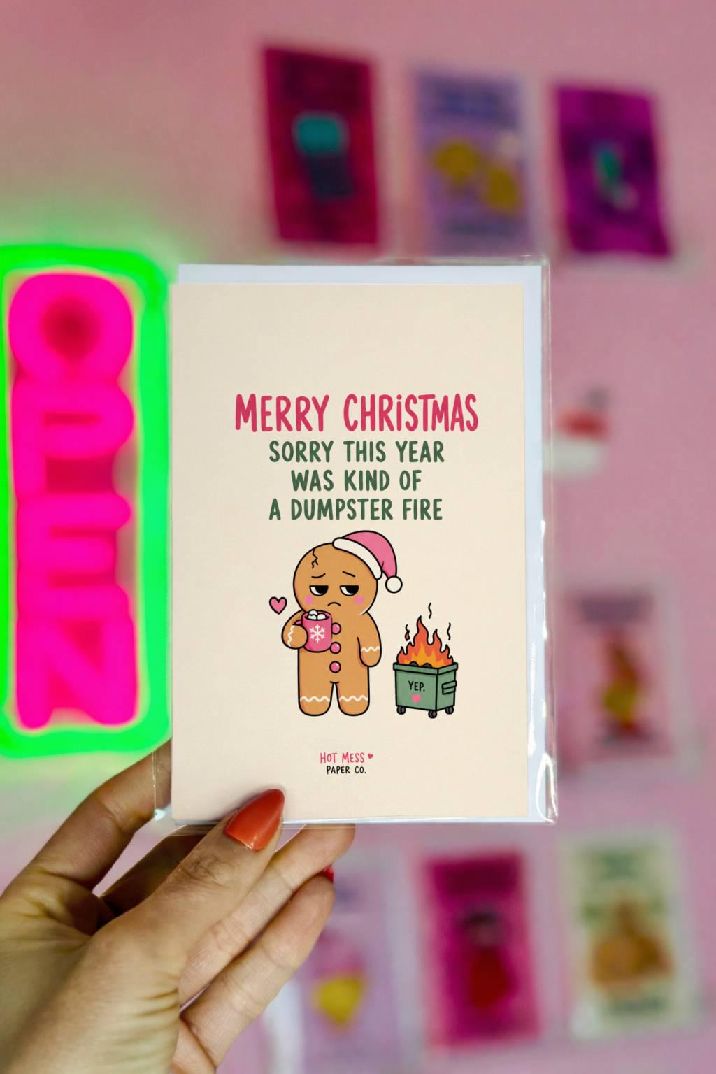 Funny Christmas card featuring a tired gingerbread cookie holding hot cocoa beside a tiny flaming dumpster with the phrase “Sorry This Year Was Kind Of A Dumpster Fire.”