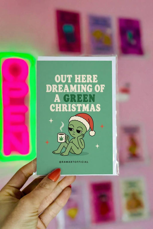 Funny Christmas card featuring an alien in a Santa hat holding a mug with a weed leaf beside the phrase “Out Here Dreaming Of A Green Christmas.”