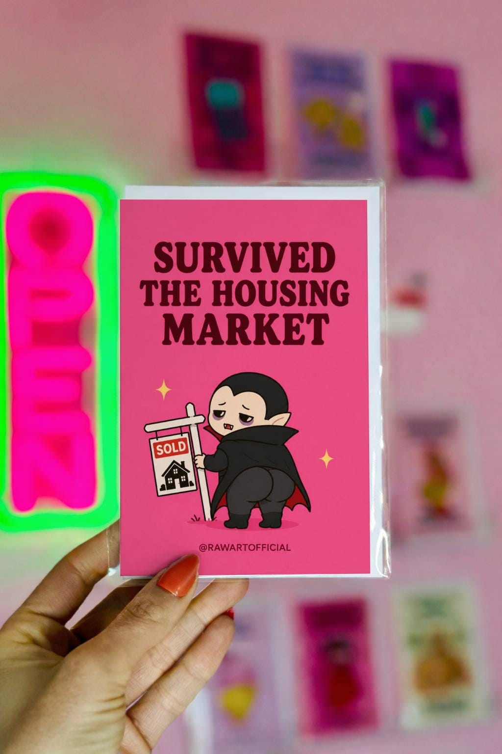 Funny new home card featuring a vampire holding a sold sign beside the phrase “Survived The Housing Market.” Humorous housewarming card for new homeowners.