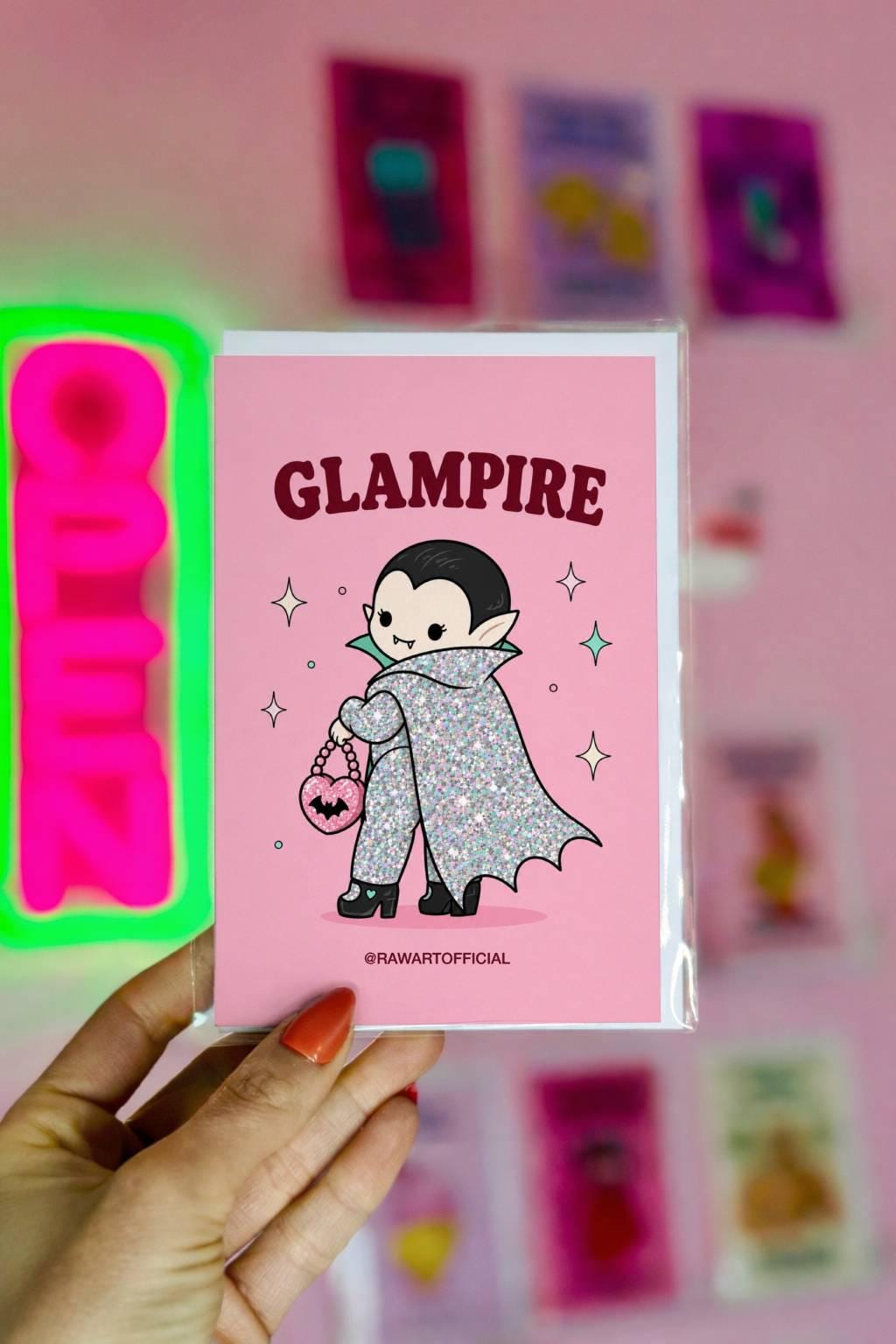 Pink Halloween greeting card reading "Glampire" featuring a cute glittery vampire wearing a glamorous cape and heels while carrying a pink heart-shaped bat purse surrounded by sparkles.