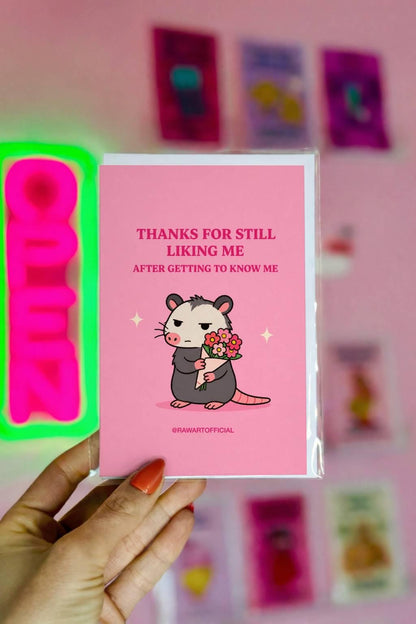 Funny greeting card featuring a grumpy opossum holding flowers beneath the phrase “Thanks for Still Liking Me After Getting to Know Me.”