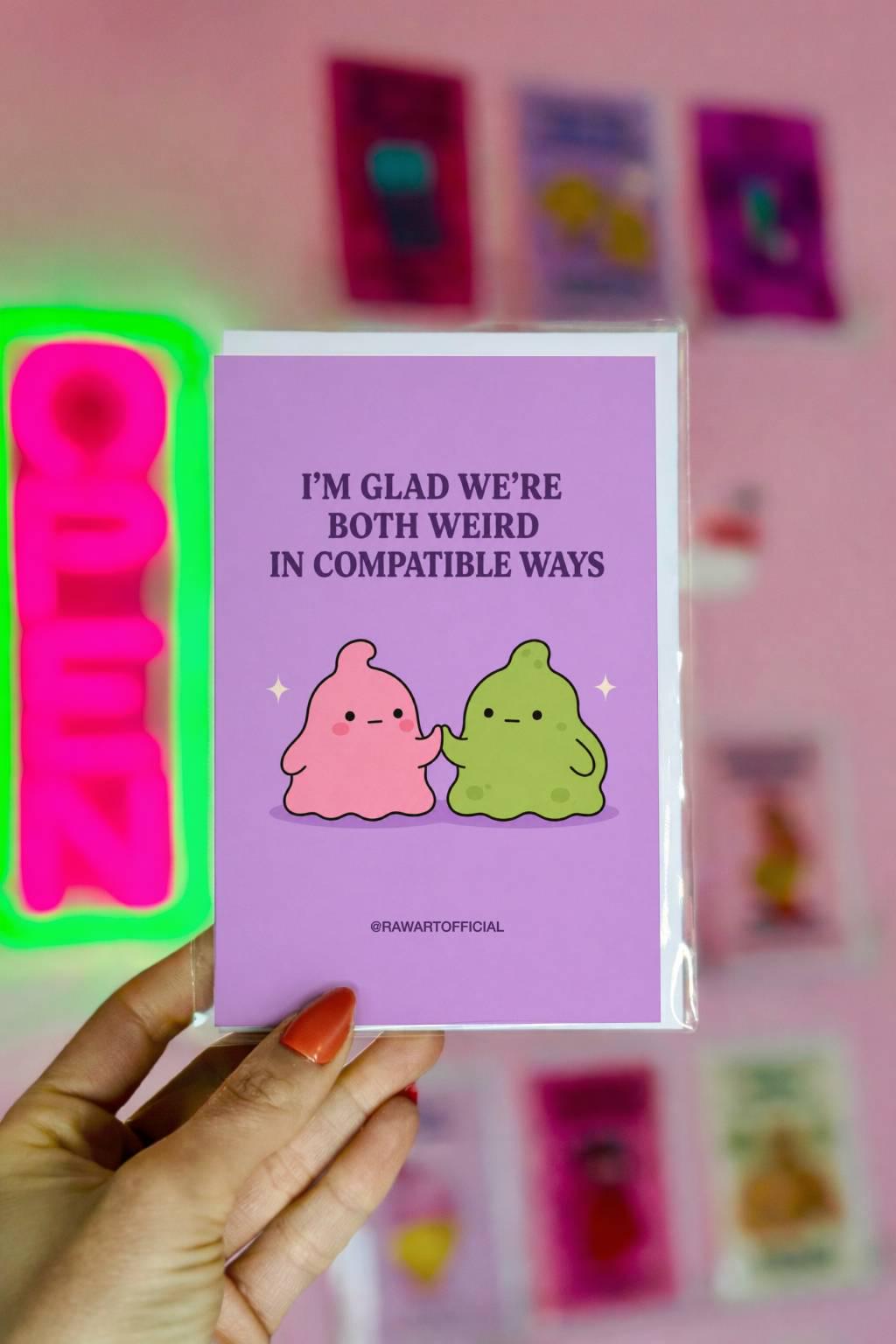 Funny love card featuring pink and green blob characters holding hands beneath the phrase “I’m Glad We’re Both Weird in Compatible Ways.”