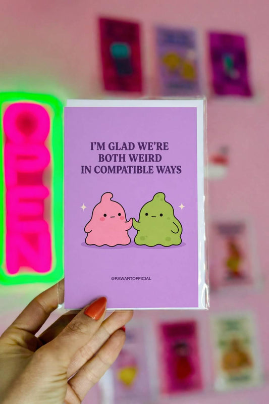 Funny love card featuring pink and green blob characters holding hands beneath the phrase “I’m Glad We’re Both Weird in Compatible Ways.”