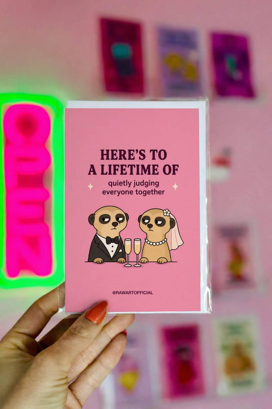 Funny wedding card featuring a bride and groom meerkat couple sharing judgmental looks over champagne with the phrase “Here’s to a Lifetime of Quietly Judging Everyone Together.”