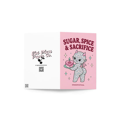 Pink funny Halloween greeting card reading “Sugar, Spice & Sacrifice” with a glittery cute devil holding a pink cake with a pentagram, photographed against a colorful pink card display.