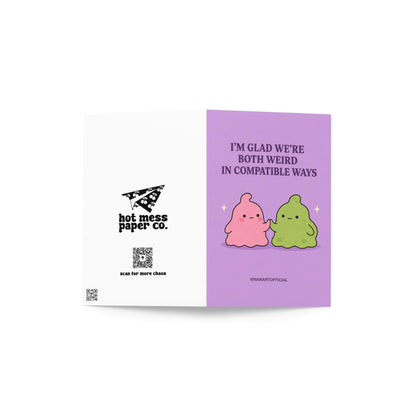 Funny love card featuring pink and green blob characters holding hands beneath the phrase “I’m Glad We’re Both Weird in Compatible Ways.”