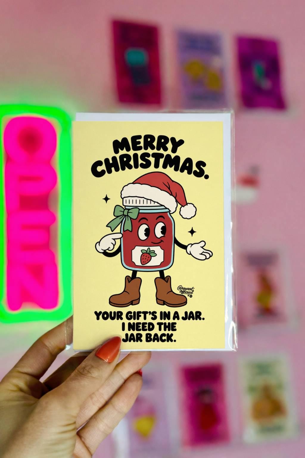 Funny Christmas greeting card featuring an illustrated strawberry jam jar character wearing a red Santa hat, green ribbon, white gloves, and brown boots on a pale yellow background. Bold black retro lettering reads “Merry Christmas. Your Gift's in a Jar. I Need the Jar Back.” Humorous holiday card inspired by homemade gifts, canning, jam making, and the importance of returning mason jars.