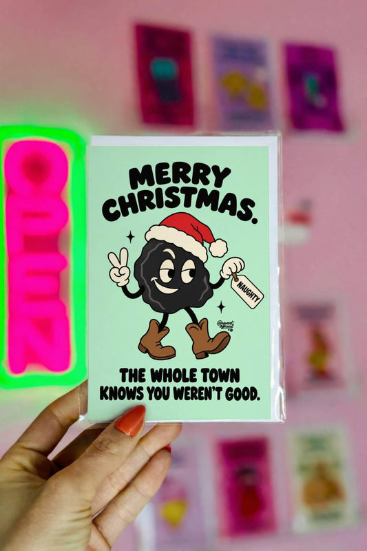 Funny Christmas greeting card featuring a smiling black lump of coal wearing a red Santa hat and brown cowboy boots, flashing a peace sign and holding a naughty list tag. Mint-green background with bold black retro lettering reading “Merry Christmas. The Whole Town Knows You Weren't Good.” Humorous small-town holiday card inspired by Christmas gossip and Santa's naughty list.