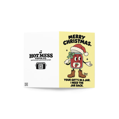 Funny Christmas greeting card featuring an illustrated strawberry jam jar character wearing a red Santa hat, green ribbon, white gloves, and brown boots on a pale yellow background. Bold black retro lettering reads “Merry Christmas. Your Gift's in a Jar. I Need the Jar Back.” Humorous holiday card inspired by homemade gifts, canning, jam making, and the importance of returning mason jars.
