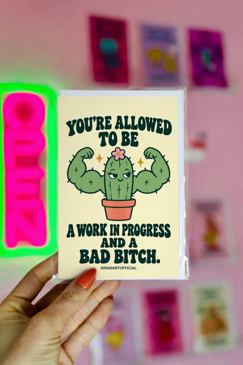Funny encouragement greeting card featuring a green cartoon cactus flexing oversized muscular arms, with a smug expression, tiny pink flower, and terracotta pot. Pale yellow background with bold dark green retro lettering reading “You're Allowed to Be a Work in Progress and a Bad Bitch.” Humorous self-love and confidence card by Hot Mess Paper Co.